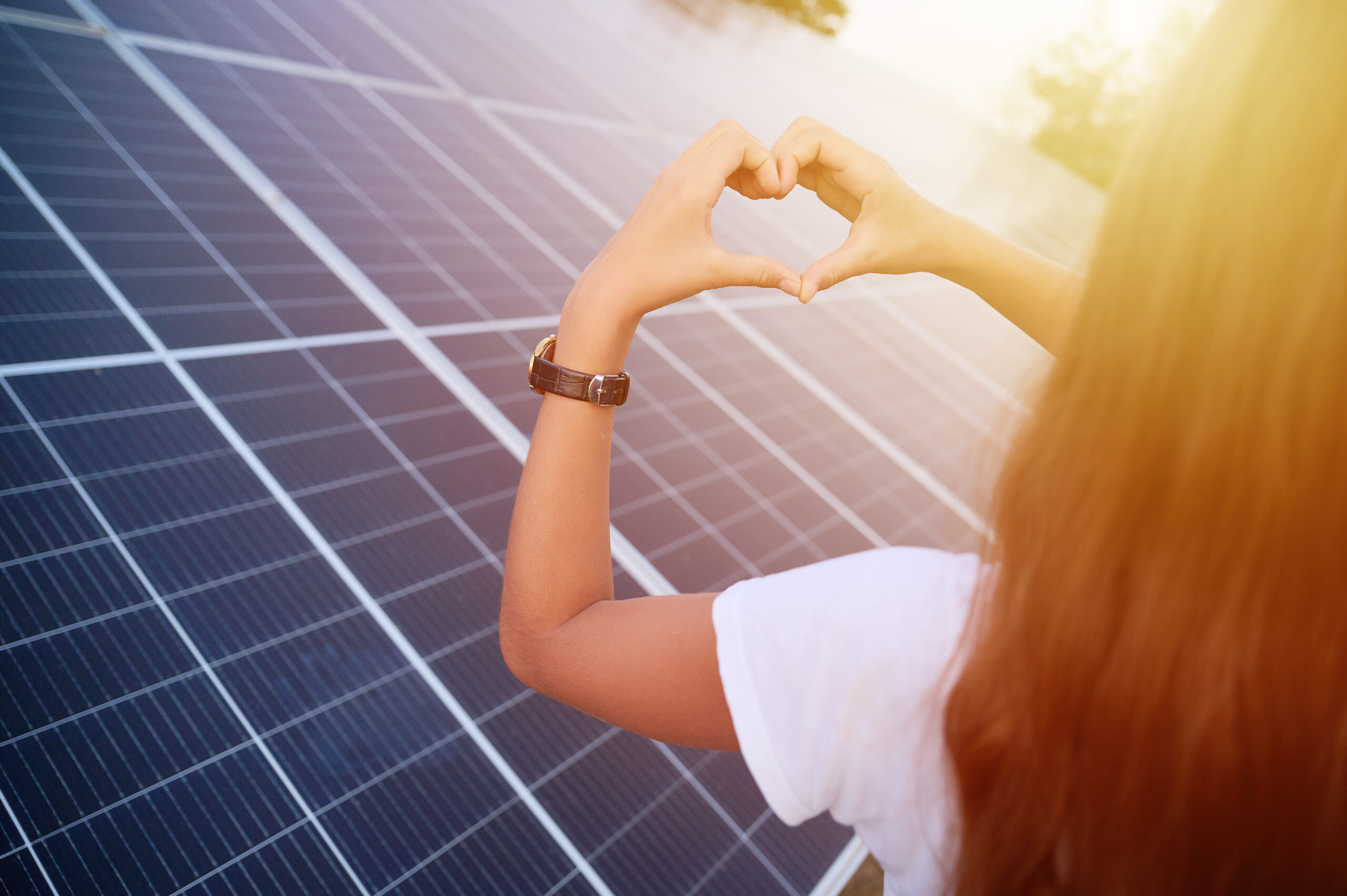 Junge Frau steht von hinten vor Solarmodulen und formt mit ihren Händen ein Herz.